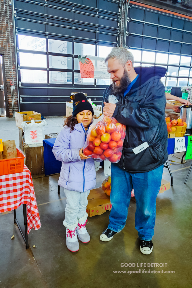 Ten Ways I'm Saving Money on Our Grocery Budget. (Photo image of my young daughter standing next to her dad and holding a large bag of apples at a farmer's market.