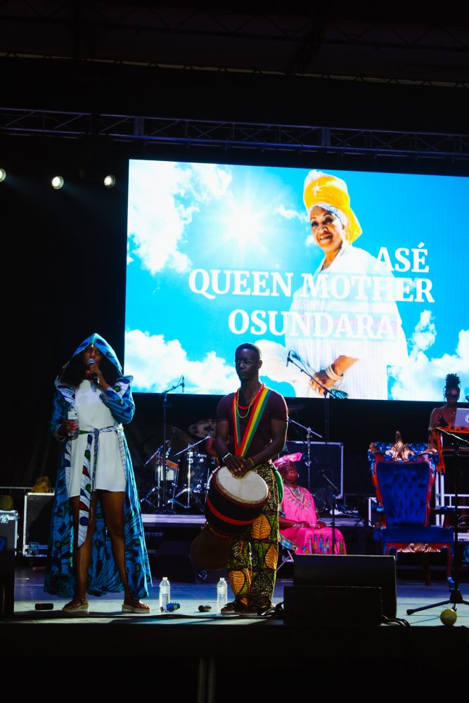 Piper Carter and Sowande Keita honor Queen Mother Osundara at the African World Festival. A photograph of Queen Mother is displayed on a large screen behind them.