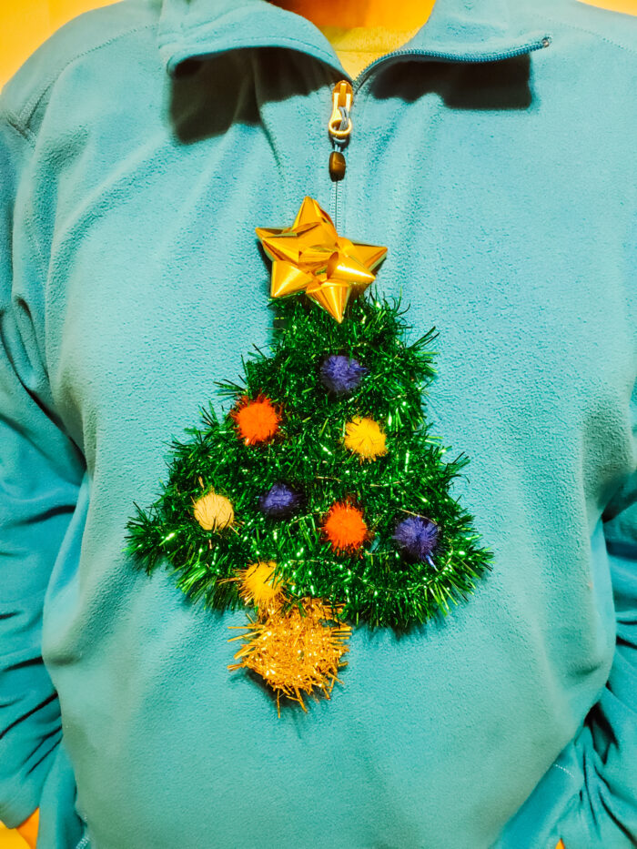 DIY Christmas Sweater Idea: a Christmas tree sewn onto a blue fleece pullover. The tree is made with green garland, a gold tree stand, and a gold gift bow to represent the star on the tree.